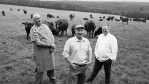 A black and white photo of a farmer, butcher and chef standing in a field surrounded by cows