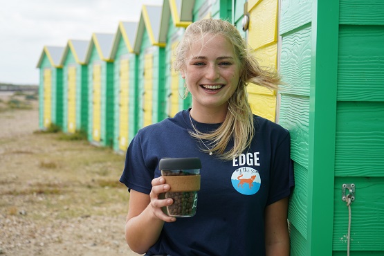 Smiling staff member from Edge by the Sea independent coffee shop infront of green and yellow beach huts in Littlehampton, Sussex